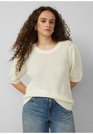 Frau mit langen lockigen Haaren, die silberne Creolen, einen strukturierten cremefarbenen Kurzarmpullover und blaue Jeans trägt, steht mit den Händen hinter dem Rücken.