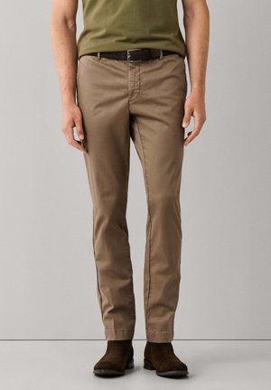 Man wearing khaki pants with a dark brown belt and dark brown suede boots standing against a plain light gray background.