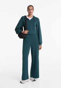 Sweat à capuche zippé teal avec capuche et manches longues, associé à un pantalon large. Porte avec un sac bandoulière noir et des sneakers de couleur claire.