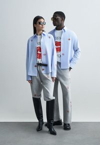 Two models stand together, wearing light blue button-up shirts with lip patterns, graphic t-shirts, grey sweatpants, and knee-high black boots.