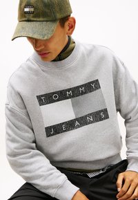 Γκρι φούτερ με το λογότυπο "TOMMY JEANS". Διαθέτει ορθογώνιο γραφικό σε μαύρο, γκρι και λευκό. Μαλακή υφή και ριμπ μανσέτες.