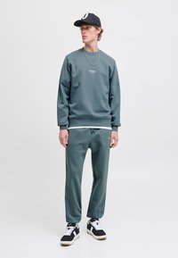 Donkergroene sweatshirt en bijpassende joggers van zacht materiaal, met een ronde hals en minimale branding. Een zwarte pet en sneakers maken de look af.