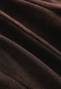 Hnedá rebrovaná tkanina s textúrovaným povrchom, ktorá obsahuje jemné línie a záhyby vytvárajúce hĺbku. Celkový vzhľad je mäkký a tlmený.
