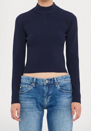 Femme portant un pull col roulé bleu marine cintré à manches longues côtelées et un jean en denim bleu clair délavé, debout les bras détendus.