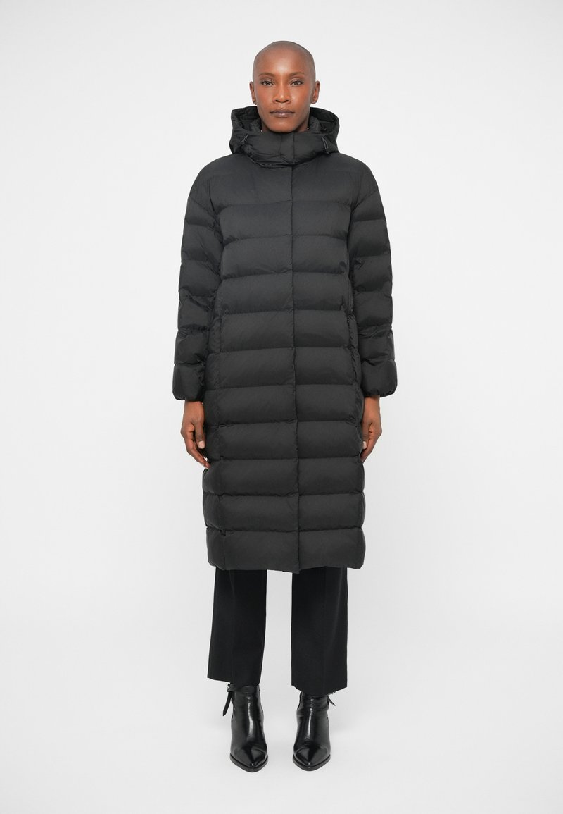 Črna puhovka z visokim ovratnikom in kapuco, z prešitim dizajnom in dolgimi rokavi, združena s črnimi hlačami in koničastimi čevlji.