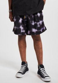 Sorte og hvite mønstrede mesh-shorts med elastisk liv, sidelommer og en lilla logo på venstre kant, sammen med svarte joggesko.