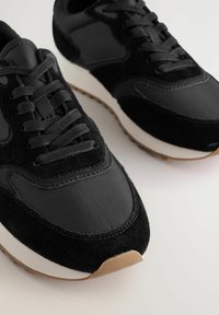 Ein Paar schwarze lässige Sneaker mit Wildleder- und Stoffakzenten, weißen Zwischensohlen und hellbraunen Gummisohlen auf einer hellen Oberfläche.