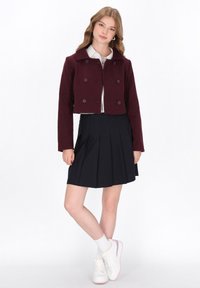 Burgundská zkrácená bunda s dvojitými knoflíky, spárovaná s skládacími námořnickými sukněmi a bílými teniskami, vytváří ležérní outfit.