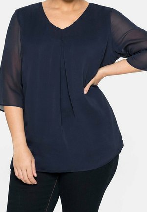 Kobieta w granatowej bluzce z przezroczystymi rękawami o długości do łokcia i plisą z przodu, w czarnych spodniach, z ręką opartą na biodrze.