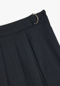 Un pantalon bleu marine à rayures dispose d'une coupe droite, de plis, et d'un accent en anneau métallique à la taille. Le matériau est lisse avec un léger brillant.