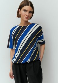 Blouse rayée bleu et blanc à manches courtes, avec des rayures horizontales et diagonales en marine, jaune et marron. Texture lisse, coupe ample.