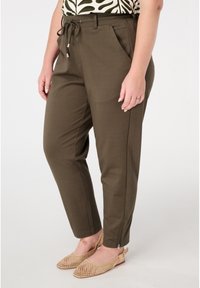 Pantalons vert olive avec taille élastique et cordon de serrage. Dotés de deux poches latérales et de zips aux chevilles. Portés avec des chaussures à talon ouvert en maille beige.