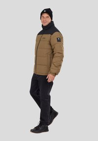 Puffer jacket in brown with black accents, featuring a high collar and zip closure. Black hat, dark jeans, and black footwear.