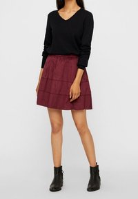 Pull noir à col en V associé à une jupe burgundy à volants, avec un tissu texturé et des bottines noires à hauteur de cheville.