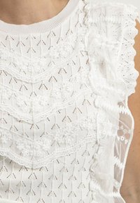 Haut sans manches blanc avec un tissu texturé orné de volants en dentelle et de motifs complexes, mettant en valeur un design en couches et des accents délicats.