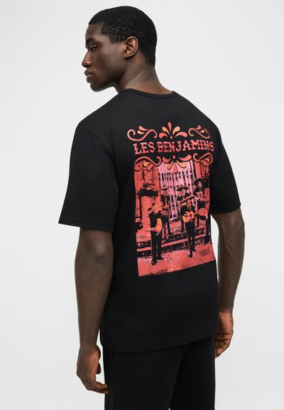 Jeune homme vu de profil portant un t-shirt noir avec un graphique rouge représentant trois musiciens et le texte "LES BENJAMINS" au dos.