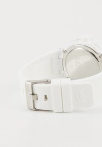 Montre de sport blanche avec un bracelet en silicone texturé, boucle en acier inoxydable poli et un fond de boîtier transparent. Détail de logo notable présent.
