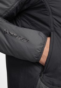 Grå isolerad jacka med en slät yta, Primaloft-logotyp på ärmen och en ficka med dragkedja på sidan.