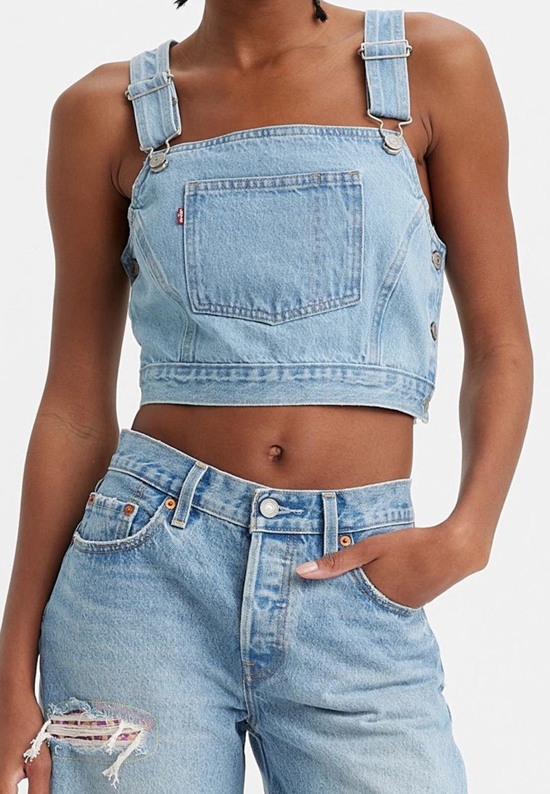 Femme portant une salopette courte en denim bleu clair avec poche avant et un jean déchiré taille haute, main dans la poche, sur fond uni.