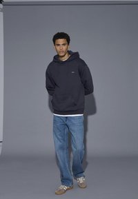 Ung mand iført en mørk hættetrøje, lyseblå jeans og beige sneakers, der står foran en ensfarvet grå baggrund.