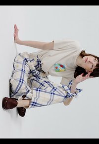 Kobieta kuca przy białej ścianie, ubrana w beżowy t-shirt z nadrukiem, szerokie spodnie w kratę z wiązanymi mankietami oraz ciemnobrązowe sandały z odkrytymi palcami.