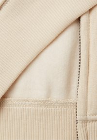 Felpa con zip beige, con texture a costine e morbido rivestimento in pile. Presenta cuciture visibili e una chiusura a zip liscia.