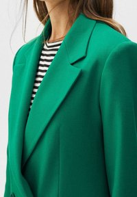 Blazer verde con un diseño ajustado, solapas marcadas y hombros estructurados, llevado sobre una blusa de rayas en blanco y negro. Textura de tela suave.