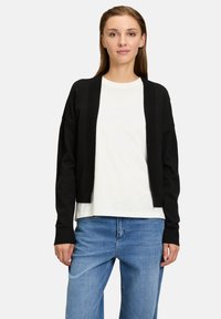 Svart stickad cardigan med öppen framsida, ribbade ärmslut och nederkant. Bärs över en vit skjorta och kombineras med ljusblå denimjeans.