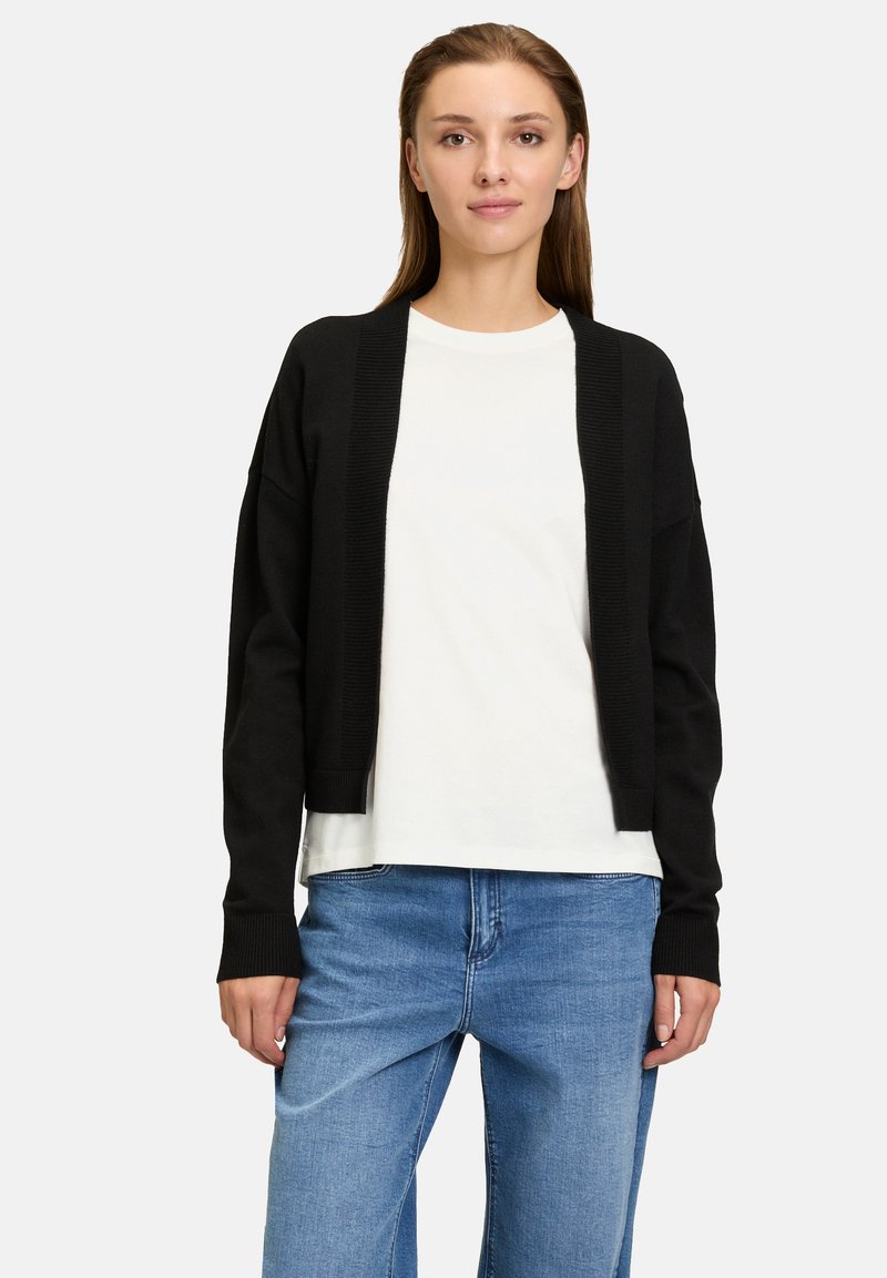 Svart stickad cardigan med öppen framsida, ribbade ärmslut och nederkant. Bärs över en vit skjorta och kombineras med ljusblå denimjeans.