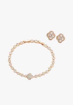 Złota bransoletka z okrągłymi przezroczystymi kamieniami i zawieszką w kształcie serca, zestawiona ze złotymi kolczykami w kształcie kwiatów, wysadzanymi małymi przezroczystymi kamieniami.