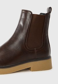 Bota de cuero marrón hasta el tobillo con paneles laterales elásticos, suela texturizada y costuras reforzadas. Cuenta con una pequeña asa en la parte posterior para facilitar su colocación.