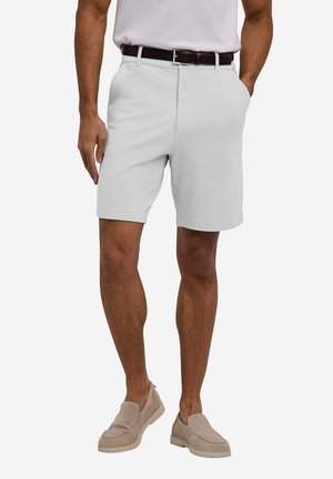 Homme portant un short gris clair mi-genou avec une ceinture marron, chemise blanche rentrée et chaussures beiges sans lacets, debout avec une main dans la poche.