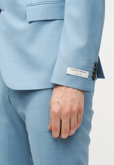 Μανίκι και παντελόνι ανοιχτό μπλε κοστουμιού με ορατή ετικέτα «Tiger of Sweden 1903» και τέσσερα σκούρα κουμπιά κοντά στη μανσέτα.
