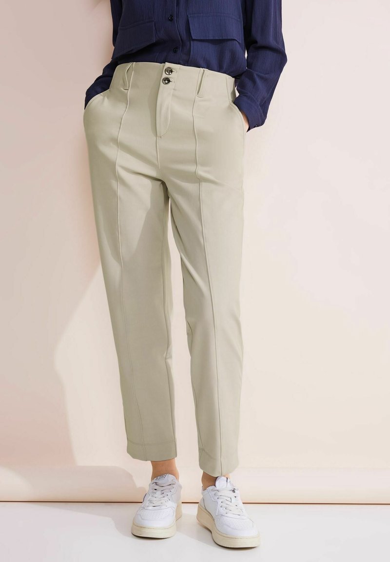 Ljusbeige skräddarsydda byxor med frontveck, två knappar och sidfickor, tillsammans med vita sneakers på en mjuk bakgrund.