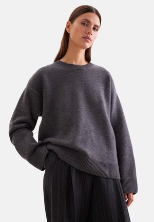 Femme portant un pull en maille gris foncé ample avec poignets et ourlet côtelés, associé à un pantalon large à fines rayures foncées, sur fond blanc.