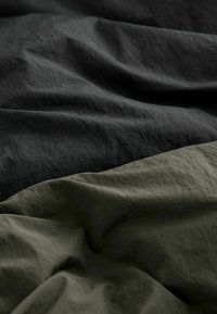 Tessuto di cotone in grigio antracite scuro e verde oliva, con una texture ondulata caratterizzata da leggere pieghe e finiture morbide e opache.