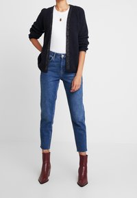 Cardigan blu navy lavorato a maglia con apertura frontale, abbinato a jeans aderenti blu e stivaletti bordeaux, caratterizzato da una finitura testurizzata e un orlo corto.