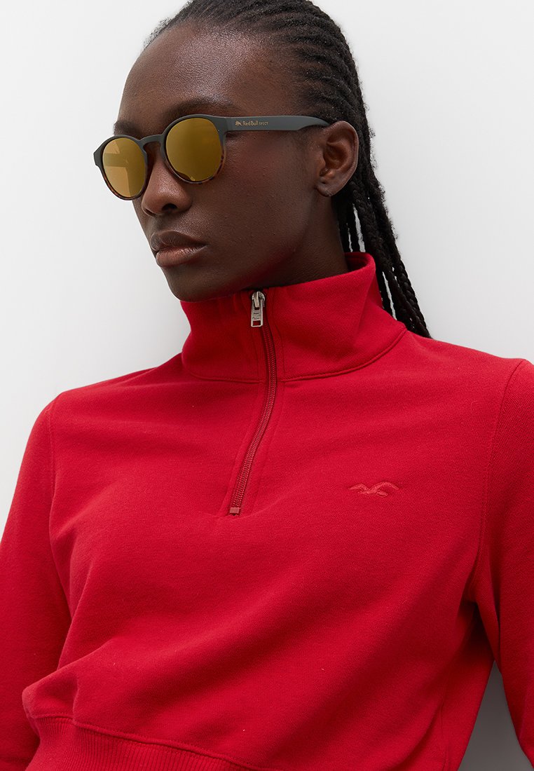 Czerwony sweatshirt z zamkiem błyskawicznym i wysokim kołnierzem, z subtelnym detalem logo. Czarne okulary przeciwsłoneczne z lustrzanymi, złotymi soczewkami o okrągłym kształcie.