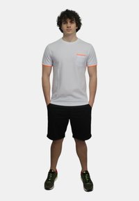 T-shirt bianco a maniche corte con rifinitura arancione e tasca. Indossato con pantaloni corti neri e sneaker verdi. Lo sfondo è bianco semplice.
