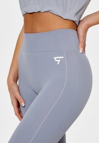 Jasnoszare legginsy z wysokim stanem wykonane z elastycznego materiału, charakteryzujące się gładką fakturą oraz białym logo po boku z kropkowanymi akcentami.