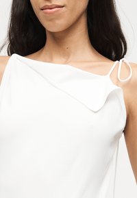 Γυναίκα που φοράει λευκό αμάνικο τοπ με ασύμμετρο ντεκολτέ και λεπτό λουρί δεμένο στον ώμο, εμφανίζοντας τον λαιμό και μερικό από το πρόσωπο.