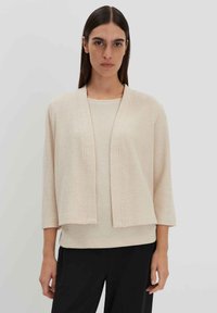 Hellbeige offene Strickjacke mit Dreiviertelärmeln, strukturiertem Stoff und Rippendetails, kombiniert mit einem passenden Oberteil und schwarzen Hosen.