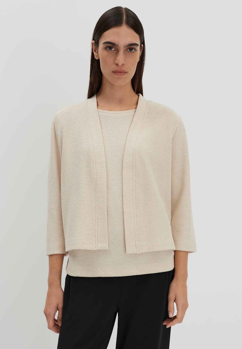 Hellbeige offene Strickjacke mit Dreiviertelärmeln, strukturiertem Stoff und Rippendetails, kombiniert mit einem passenden Oberteil und schwarzen Hosen.