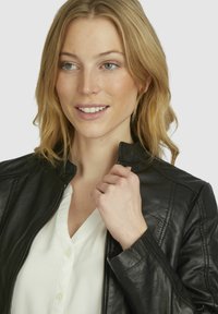 Black leather jacket with a high collar, zip closure, and visible stitching details, worn over a white button-up shirt.