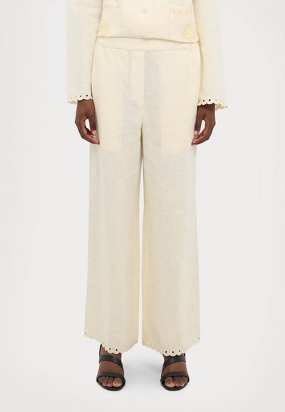 Pantalons en lin à jambe large couleur crème, avec un ourlet festonné et des découpes, et une texture lisse. Portés avec des sandales noires à lanières.