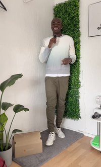 Hvid til lysegrå gradient sweater, olivengrøn joggerbukser, hvide sneakers. Indendørs omgivelser med en potteplante og en papkasse.