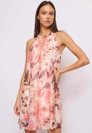 Giovane donna con abito senza maniche a fiori rosa, stile halter, che posa con una mano sul fianco davanti a uno sfondo neutro.