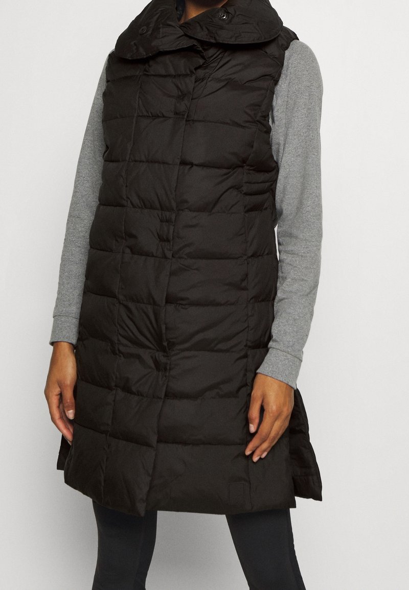 Femme portant une veste matelassée noire sans manches sur un haut gris à manches longues et un pantalon noir, debout devant un fond uni.
