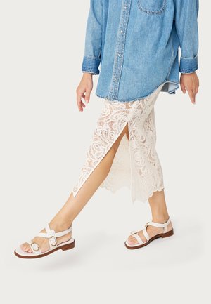 Mujer caminando con una falda blanca de encaje con abertura lateral, camisa de mezclilla azul claro y sandalias blancas con tira y hebilla sobre un fondo blanco.