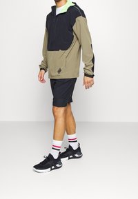 Kevyt, vetoketjullinen pullover-takki oliivivihreänä ja mustana, limenvihreällä hupulla, yhdistettynä mustiin urheilushortseihin ja mustiin lenkkareihin.